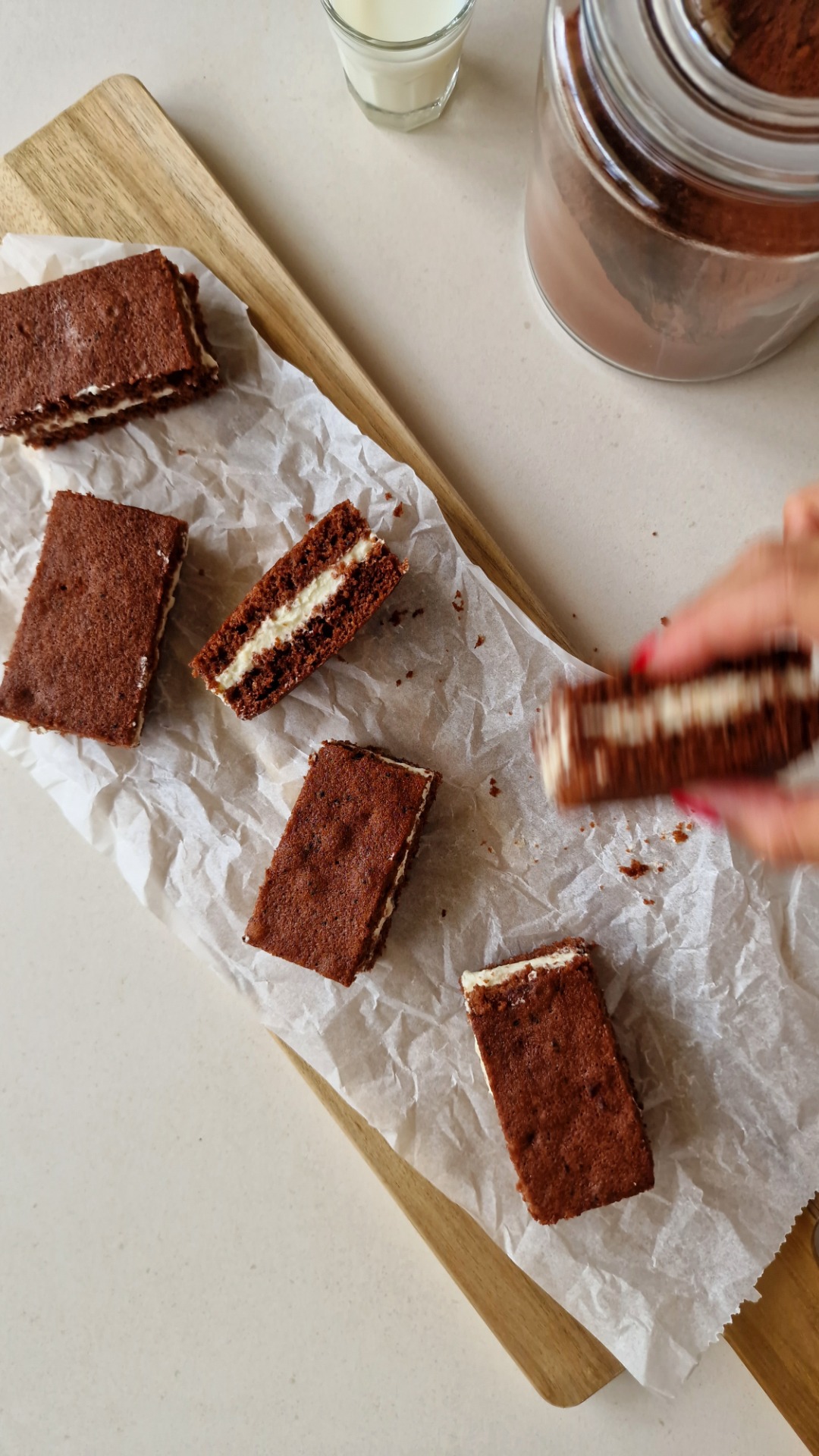 Milchschnitte auf Backpapier, daneben Glas Milch und Glas mit Kakaopulver im Hintergrund.
